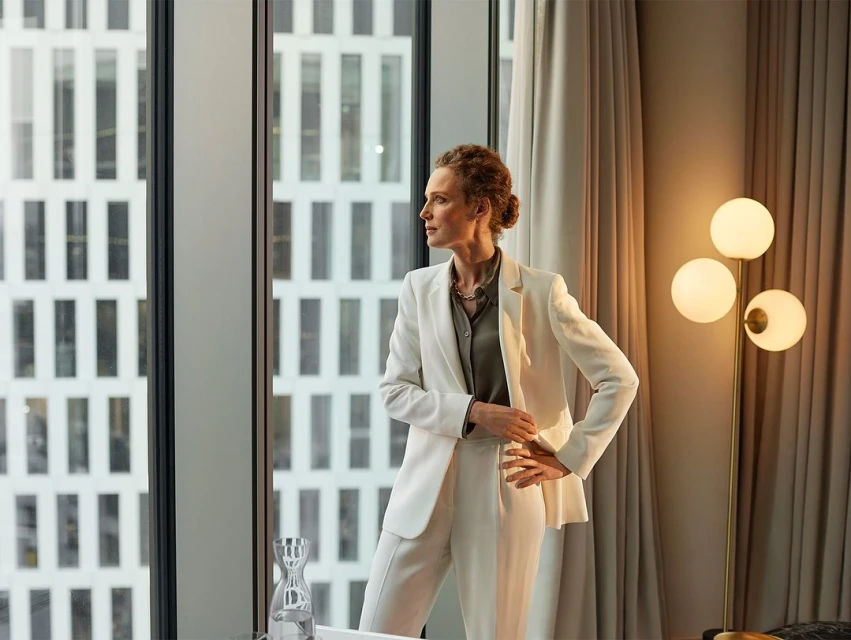 Elegant woman in white suit looks out window in modern office. Light from large windows brightens the room with modern decor.