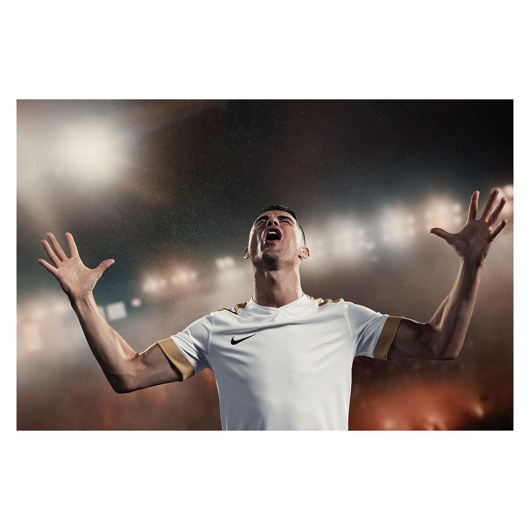 A soccer player in uniform stands on a field with arms raised. He has short, dark hair. Stadium lights illuminate the background.