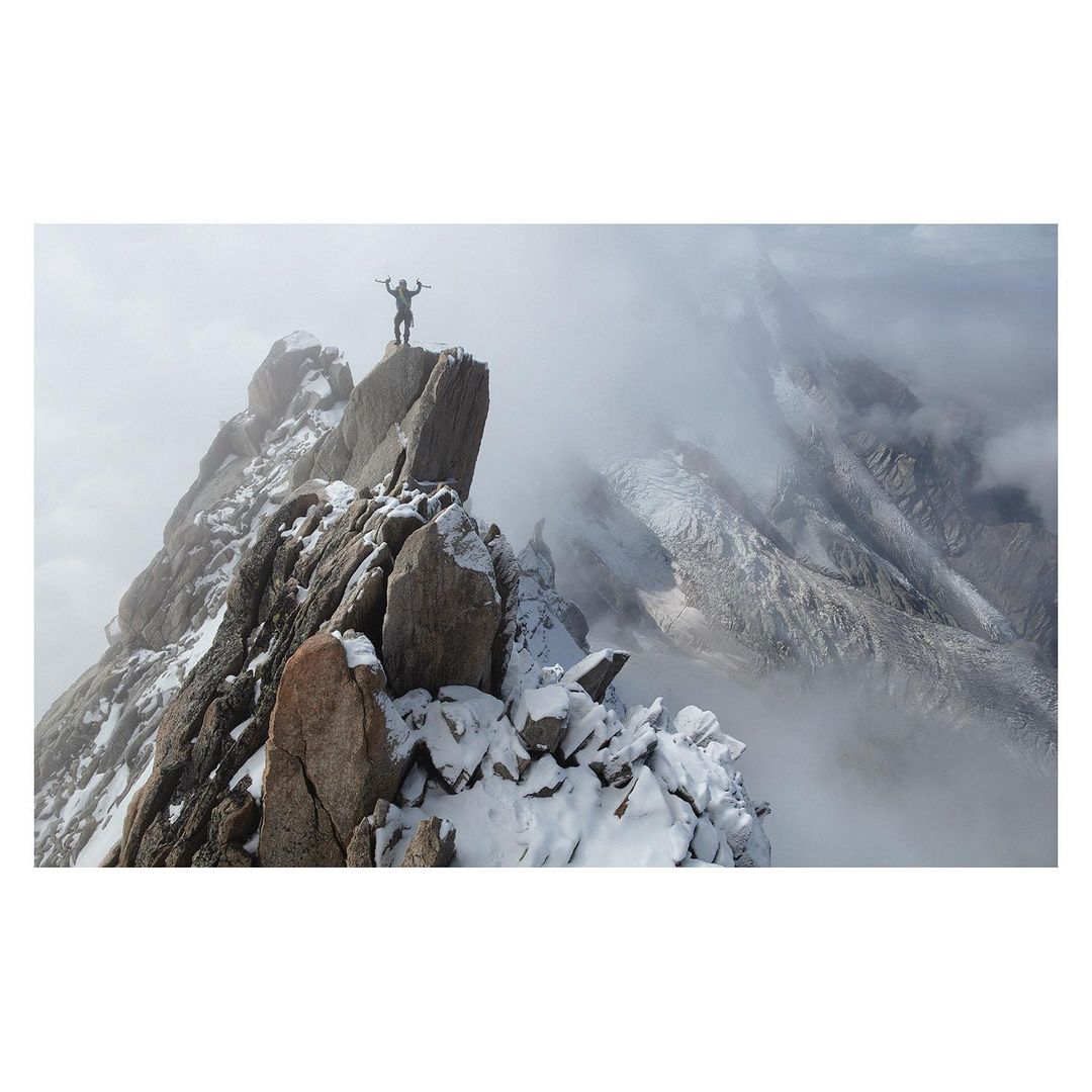 Silhouette of a person with arms raised atop a snowy, rocky mountain peak, surrounded by clouds and a hazy mountain range in the background.