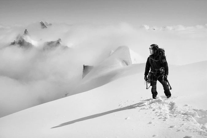 Black and white shot of a mountaineer on a snowy peak with mountains in the background and cloud cover, wearing cold-weather gear.