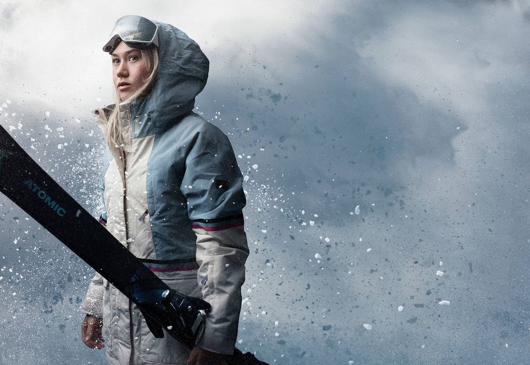 Woman in ski gear and goggles poses outdoors with ski against a blue and white snowy background with some motion blur; winter sport.
