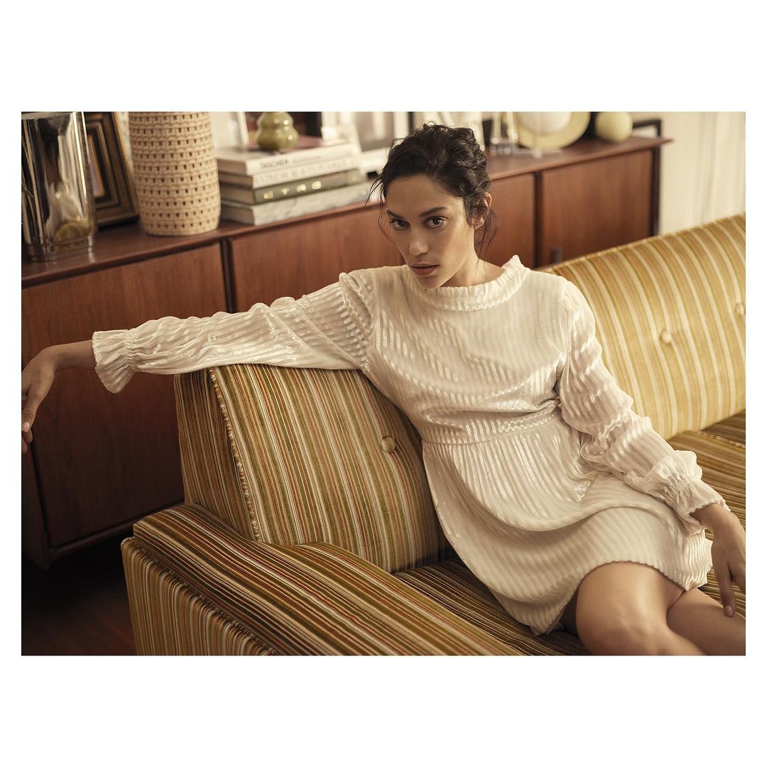 Elegant woman in a white textured dress poses on a striped couch in an indoor setting with warm lighting, creating a fashion forward look.