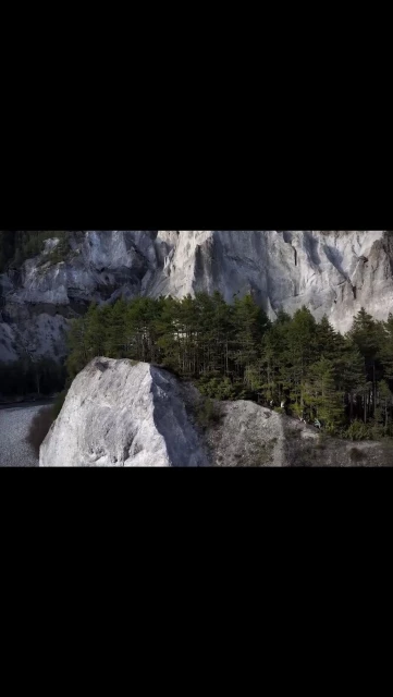 Scenic view of a rock formation with trees on top, with a rugged cliff face and a river visible in the background on a bright day.