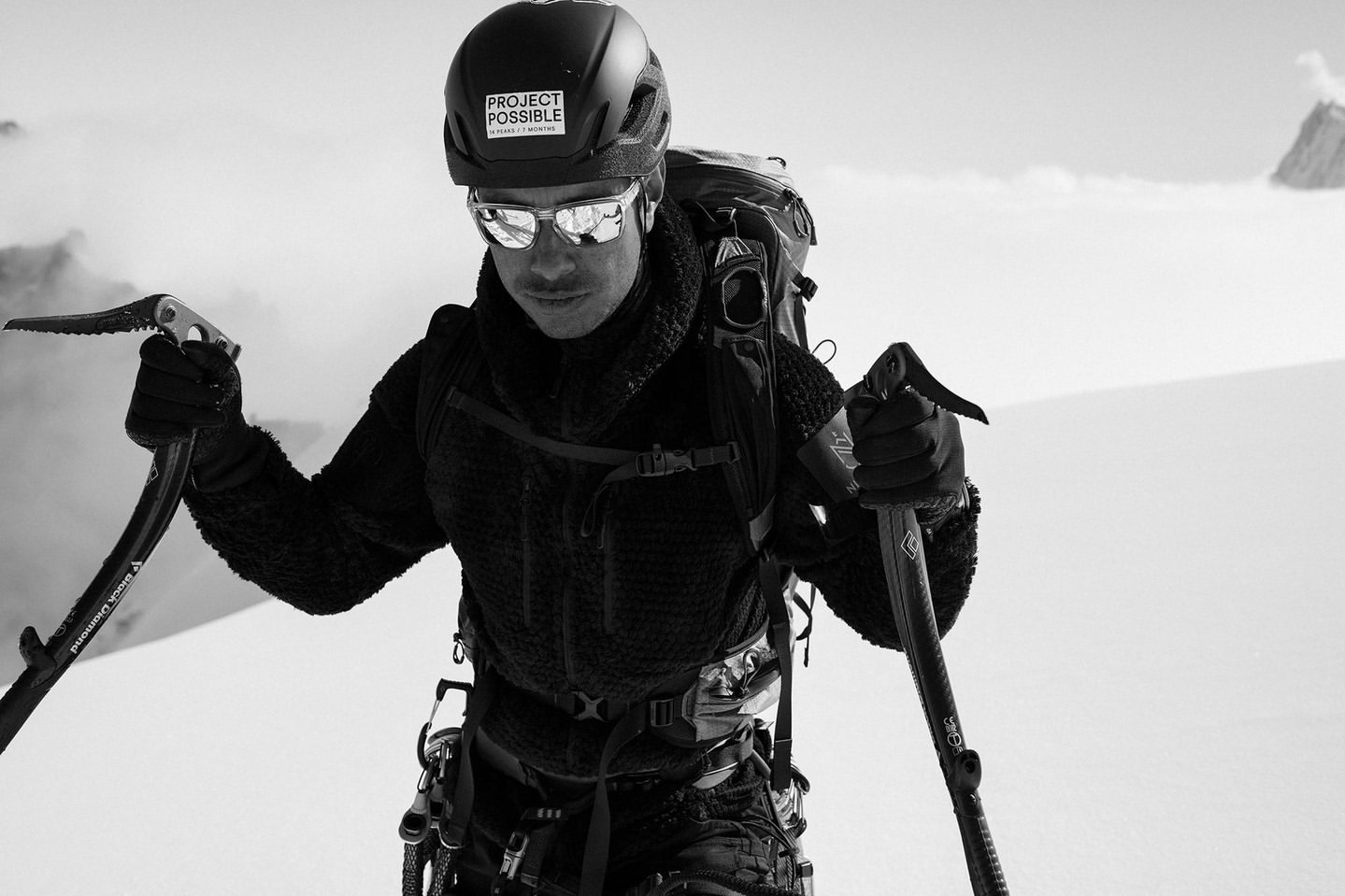 Monochrome shot of a mountaineer wearing a helmet and sunglasses while hiking on a snowy mountain, with ice axes in both hands.