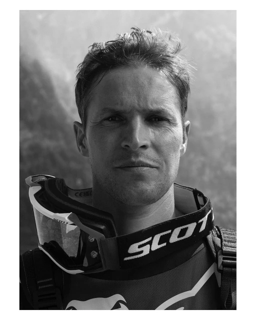 Monochromatic portrait of an adult male athlete wearing sporting goggles and a neck brace against a blurred mountain background.