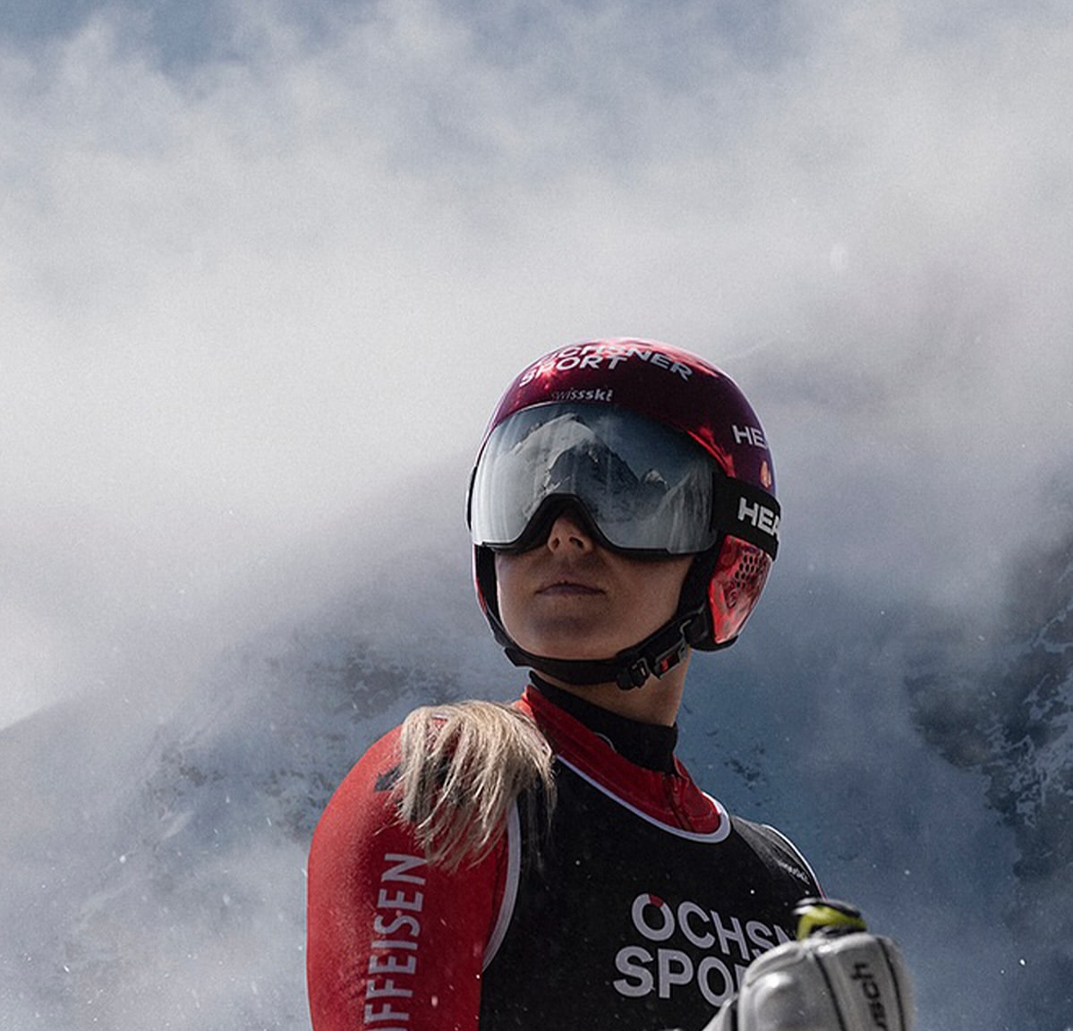 Woman skier in red and black ski suit with helmet and goggles, looking up, in a snowy mountain environment.
