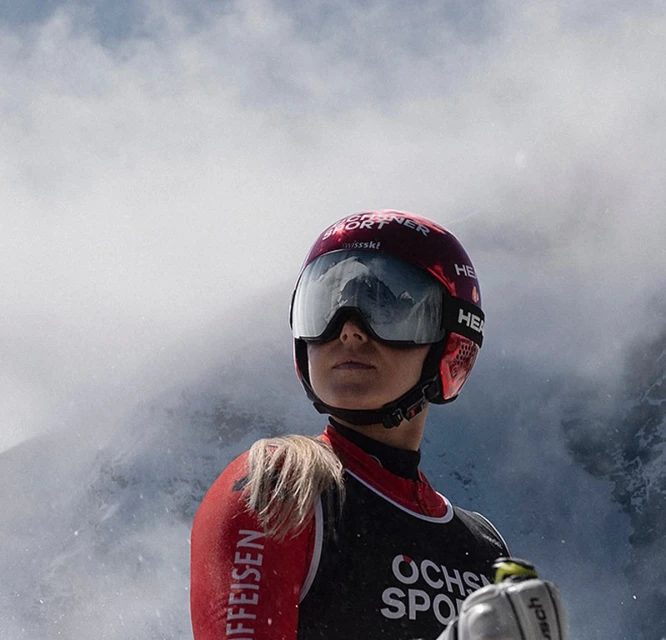 Woman skier in red and black ski suit with helmet and goggles, looking up, in a snowy mountain environment.