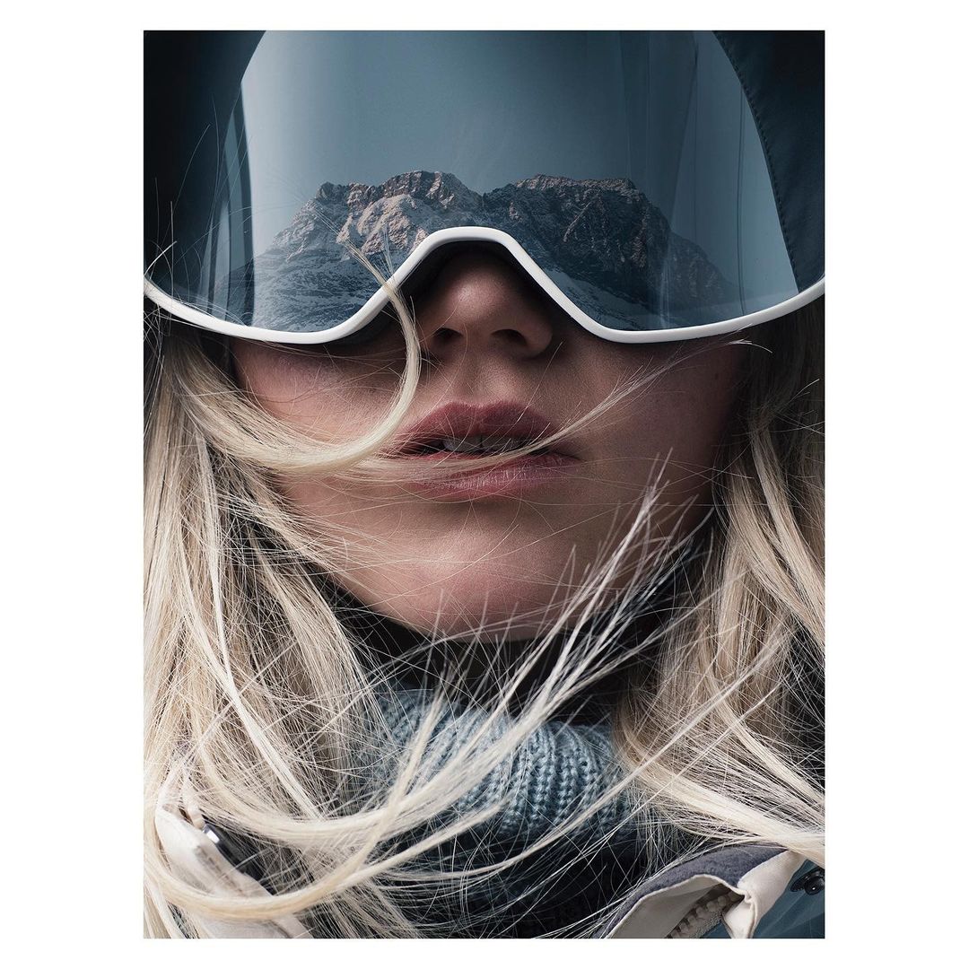 Close-up of a woman with blond hair wearing ski goggles reflecting a mountain range, portraying winter sports and outdoor adventures.