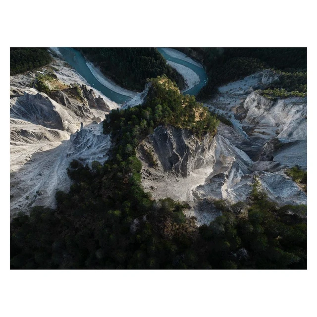 Aerial view of the confluence of a blue river with a grey rocky mountain landscape covered in green trees and vegetation.