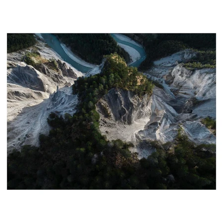 Aerial view of the confluence of a blue river with a grey rocky mountain landscape covered in green trees and vegetation.