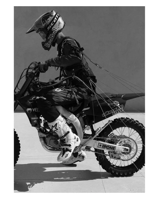 Black and white action shot of a motocross rider on a dirt bike with full safety gear, helmet, and racing suit against a plain background.