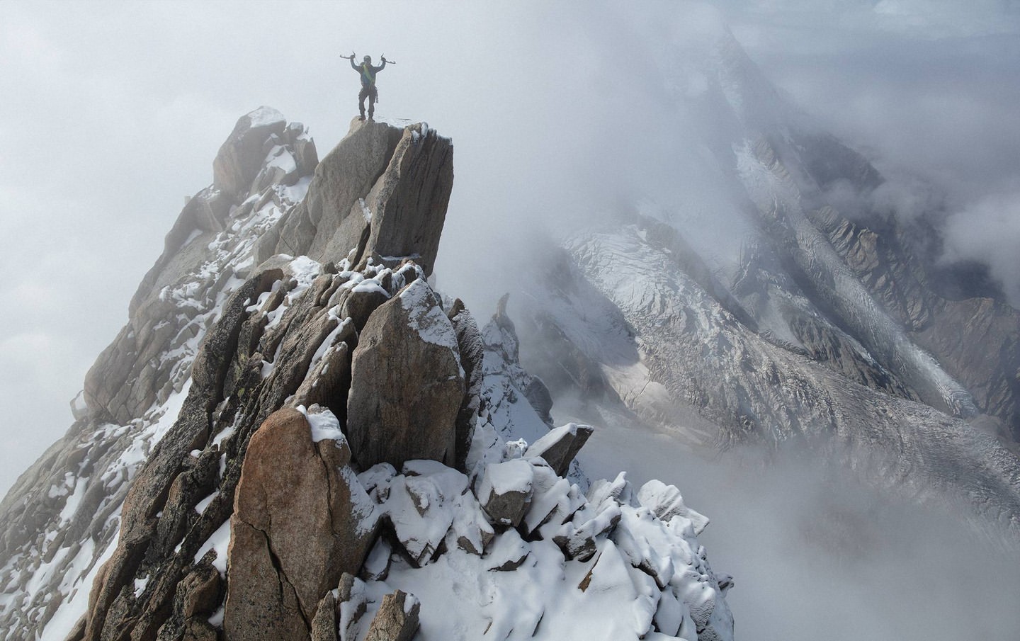 A silhouetted adult figure stands triumphantly atop a rocky, snow-dusted mountain peak, shrouded in mist, with towering snow-covered mountains in the distance.