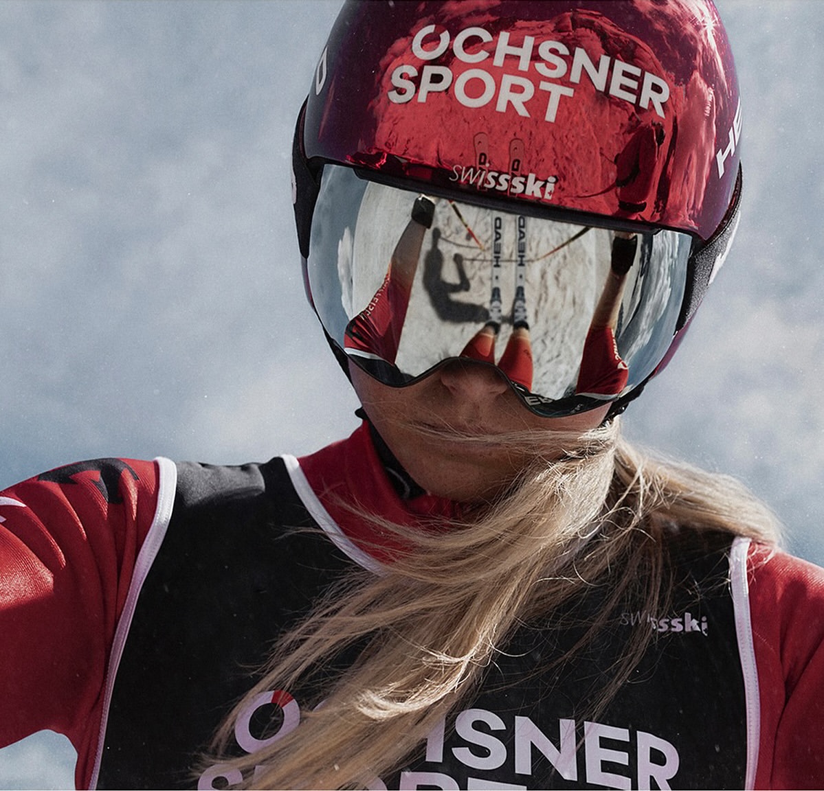 Close-up of a female skier in red and black outfit with a helmet and goggles reflecting the snowy mountain, against a cloudy sky.