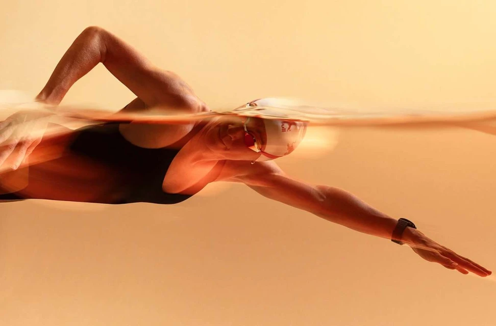 Underwater shot of a swimmer with goggles and a cap in action, showing their arm extended forward, set against a neutral background.