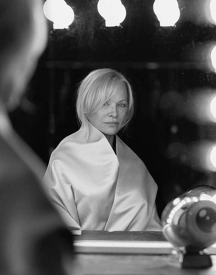 Black and white portrait of a blond woman in a satin wrap, looking into a mirror illuminated by vanity lights, reflecting back at the viewer.