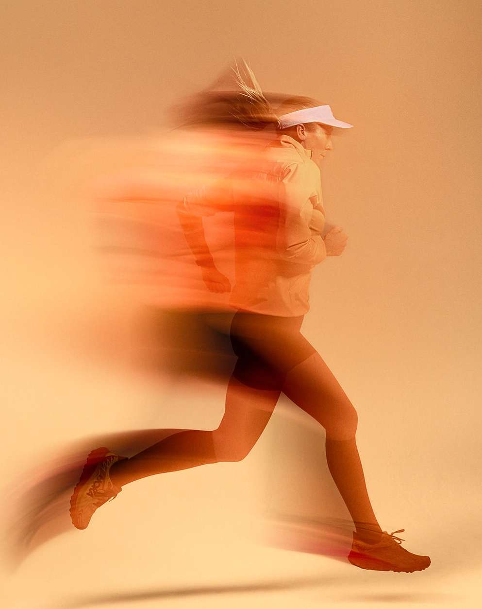 Motion blur image of adult female runner in visor and jacket against solid background, creating a sense of speed and energy.