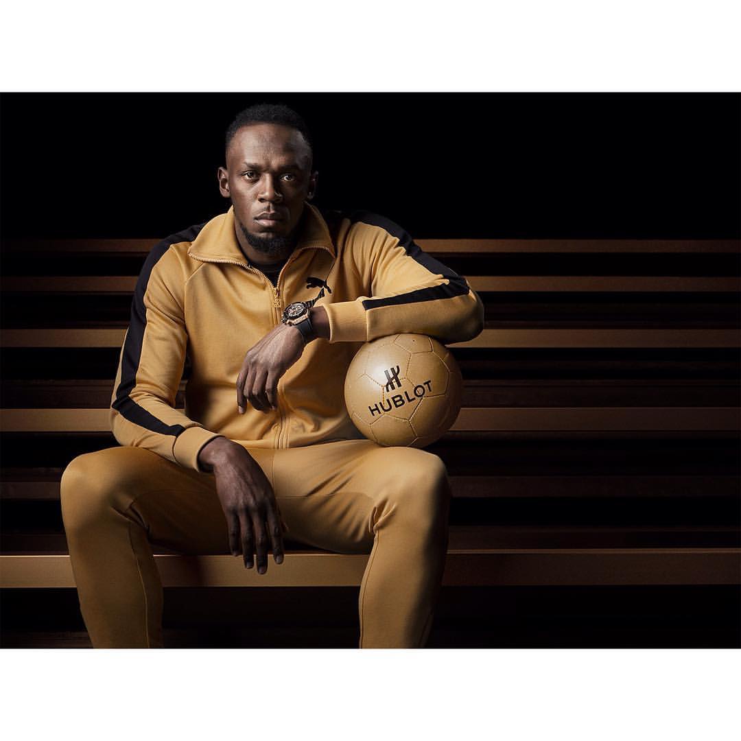 Usain Bolt, athletic man in gold tracksuit, sits with soccer ball indoors on steps; athletic fashion, sponsored by Hublot.