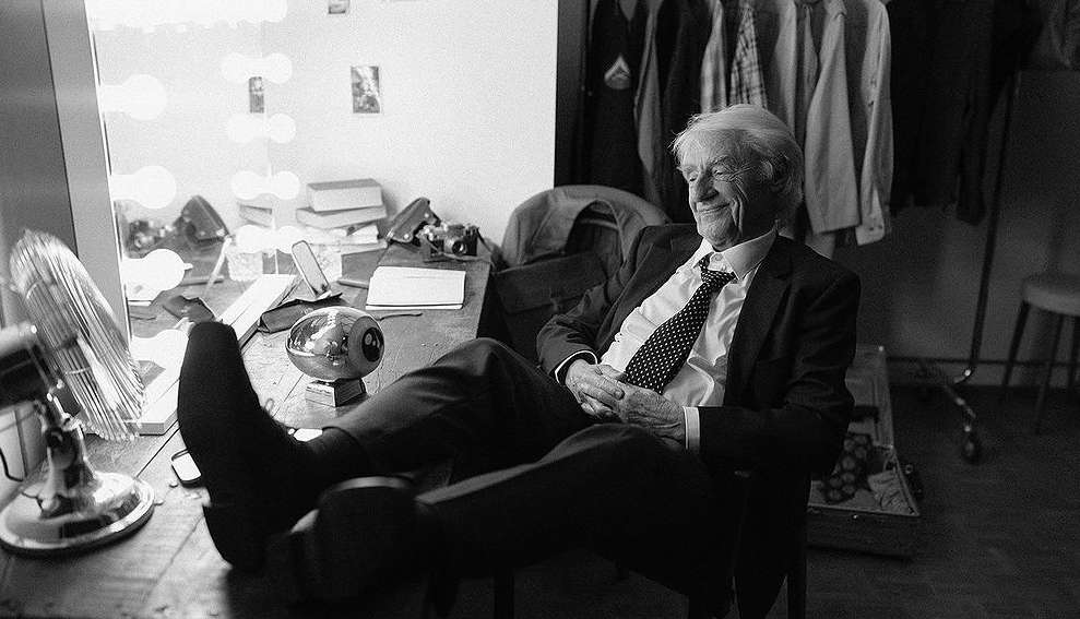Black and white photo of an elderly man in a suit relaxing at a desk with a vintage fan and other props, exuding a calm and confident vibe.