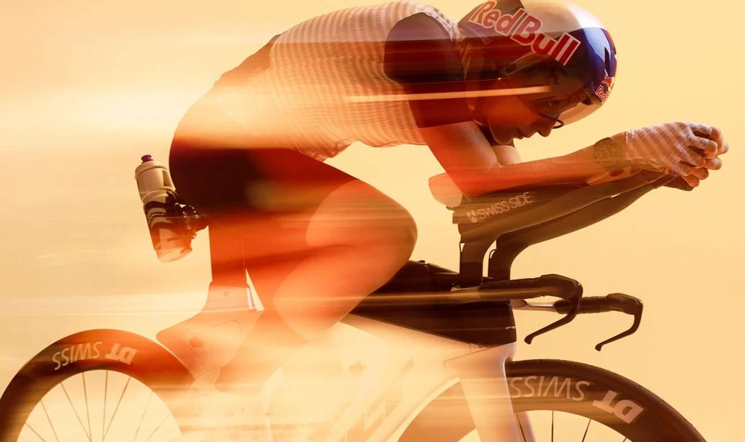 A woman wearing a helmet and racing gear rides a bike in a time trial against an orange background, showing movement with motion blur.