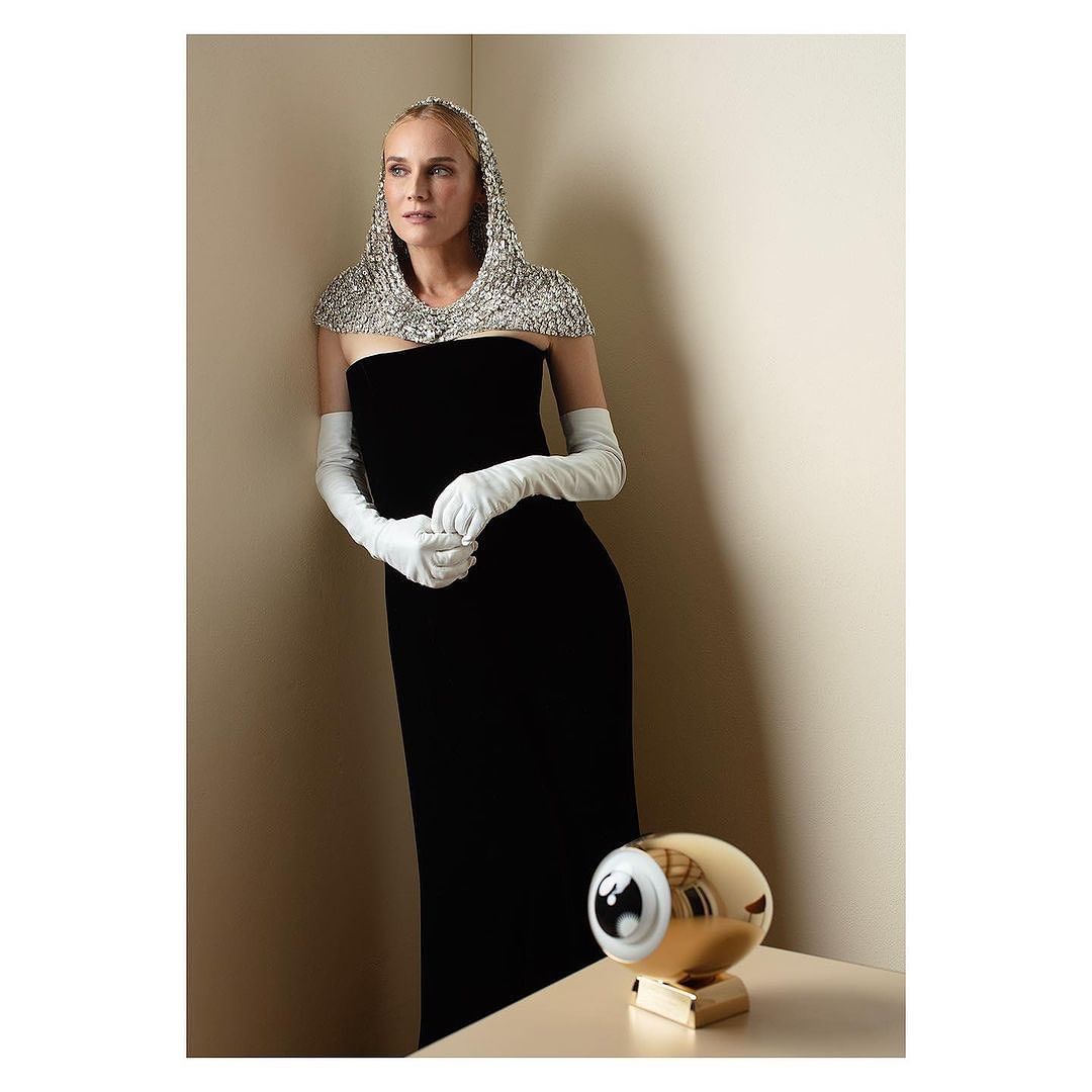 Elegant woman in a black strapless dress and white gloves poses in front of a tan wall with a decorative gold robot lamp.