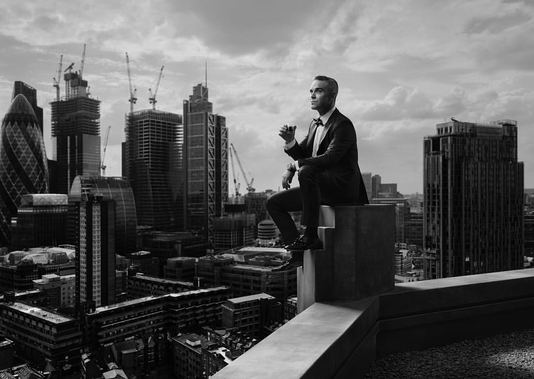 Monochrome portrait of a man in a suit sitting on a rooftop overlooking a city skyline with skyscrapers and construction cranes.