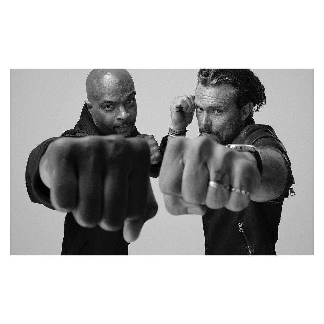 Black and white studio shot of two adult men in a dynamic pose with their fists up, wearing dark clothing, against a light backdrop.
