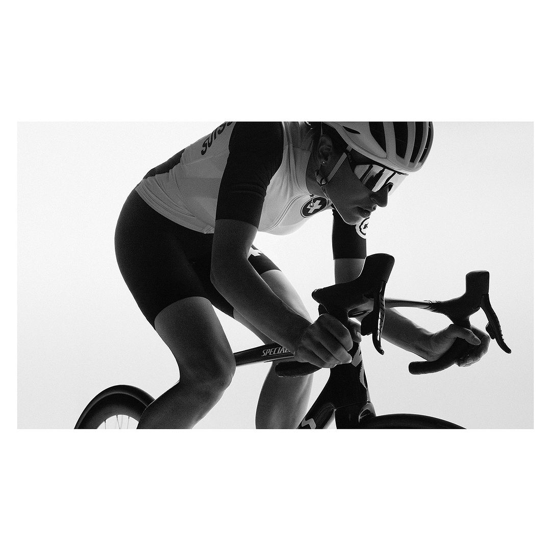 Black and white studio shot of a woman riding a Specialized racing bicycle with aerodynamic helmet and sunglasses.
