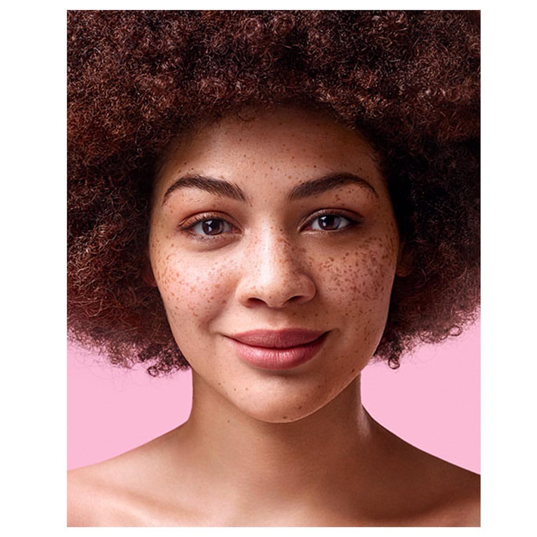 Close-up portrait of a woman with freckles and voluminous dark curly hair against a pink background