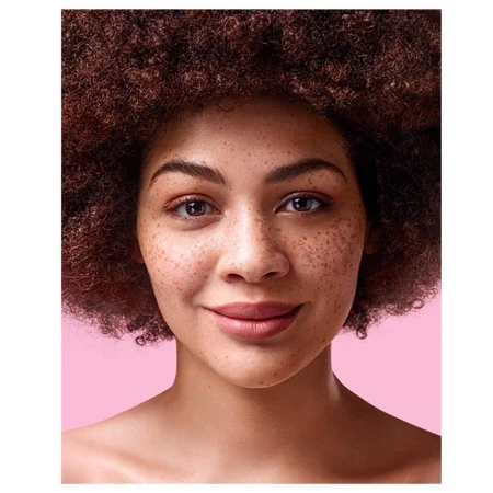 Close-up portrait of a woman with freckles and voluminous dark curly hair against a pink background