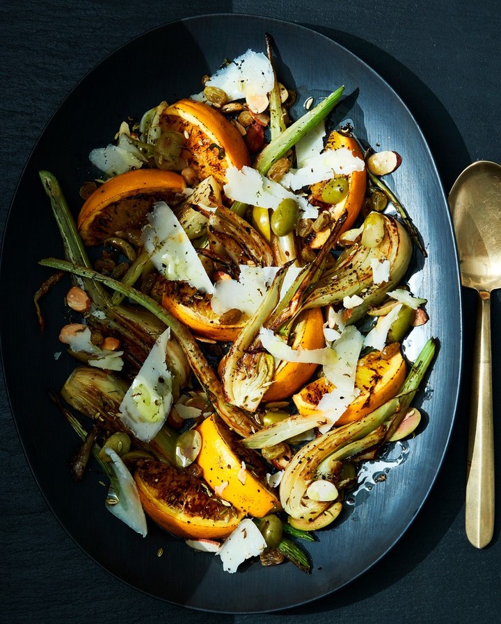 Overhead shot of a dark plate featuring a salad of oranges, fennel, olives, green beans, and shaved cheese, beside a gold spoon.