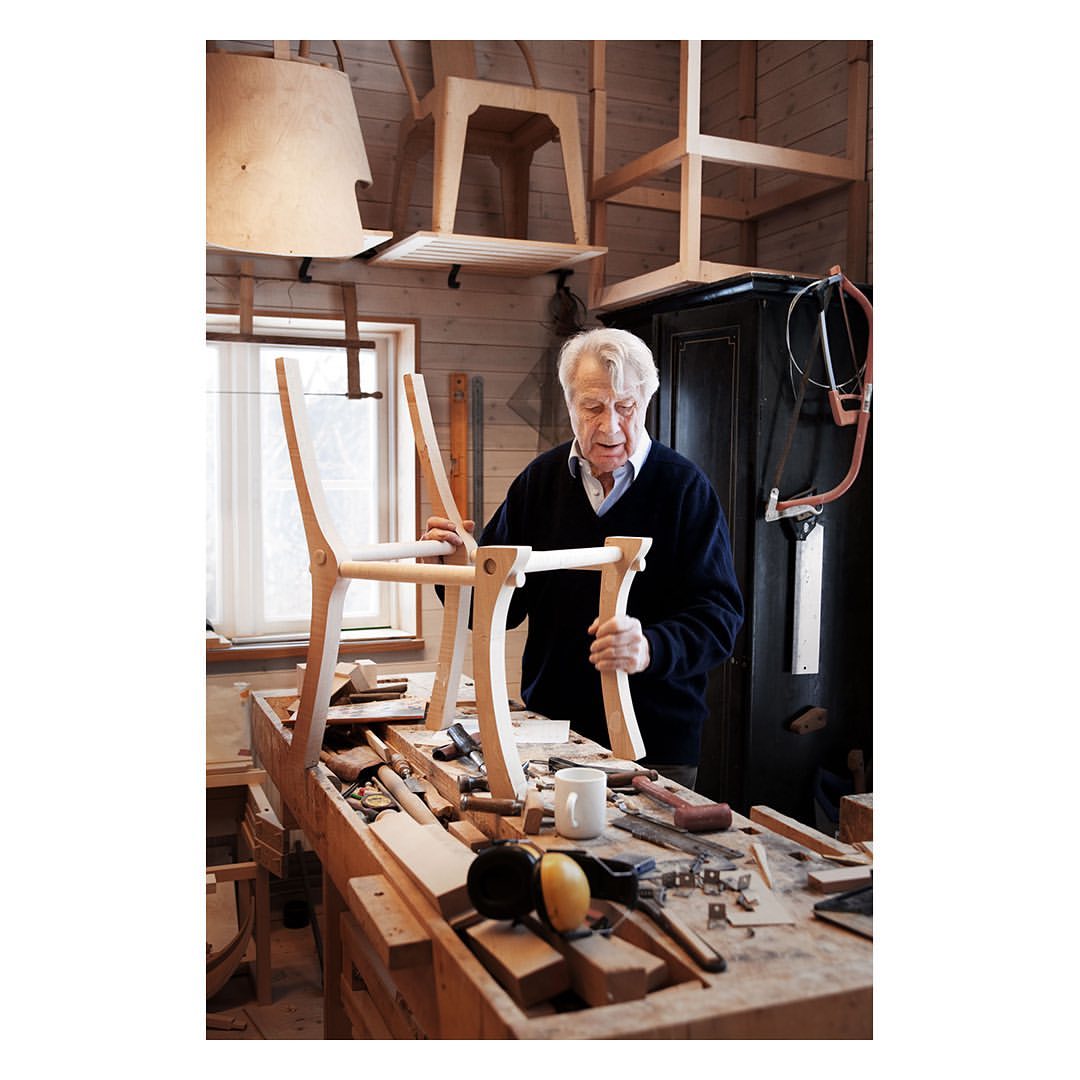 Elderly man assembling a wooden chair frame at a workbench in his workshop, surrounded by tools and other chair parts.