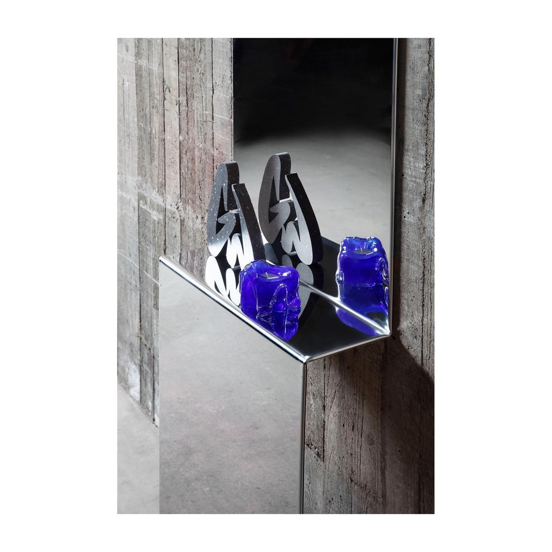Abstract mirrored installation with blue glass skulls on shelf, next to a grey concrete wall in a dark room setting indoors.
