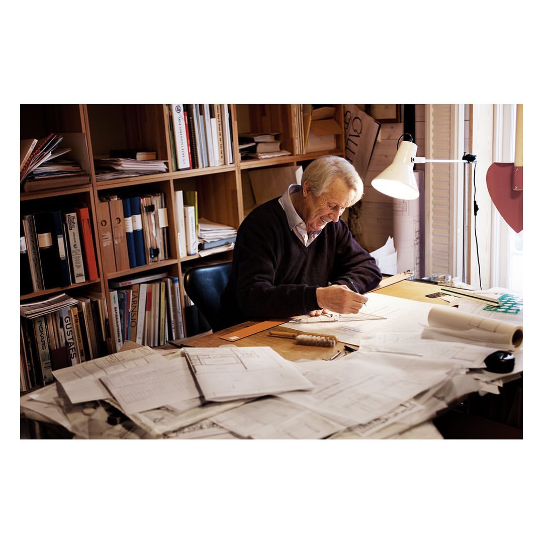 Senior man smiles as he draws at a table cluttered with architectural plans in a room filled with bookshelves and natural light.