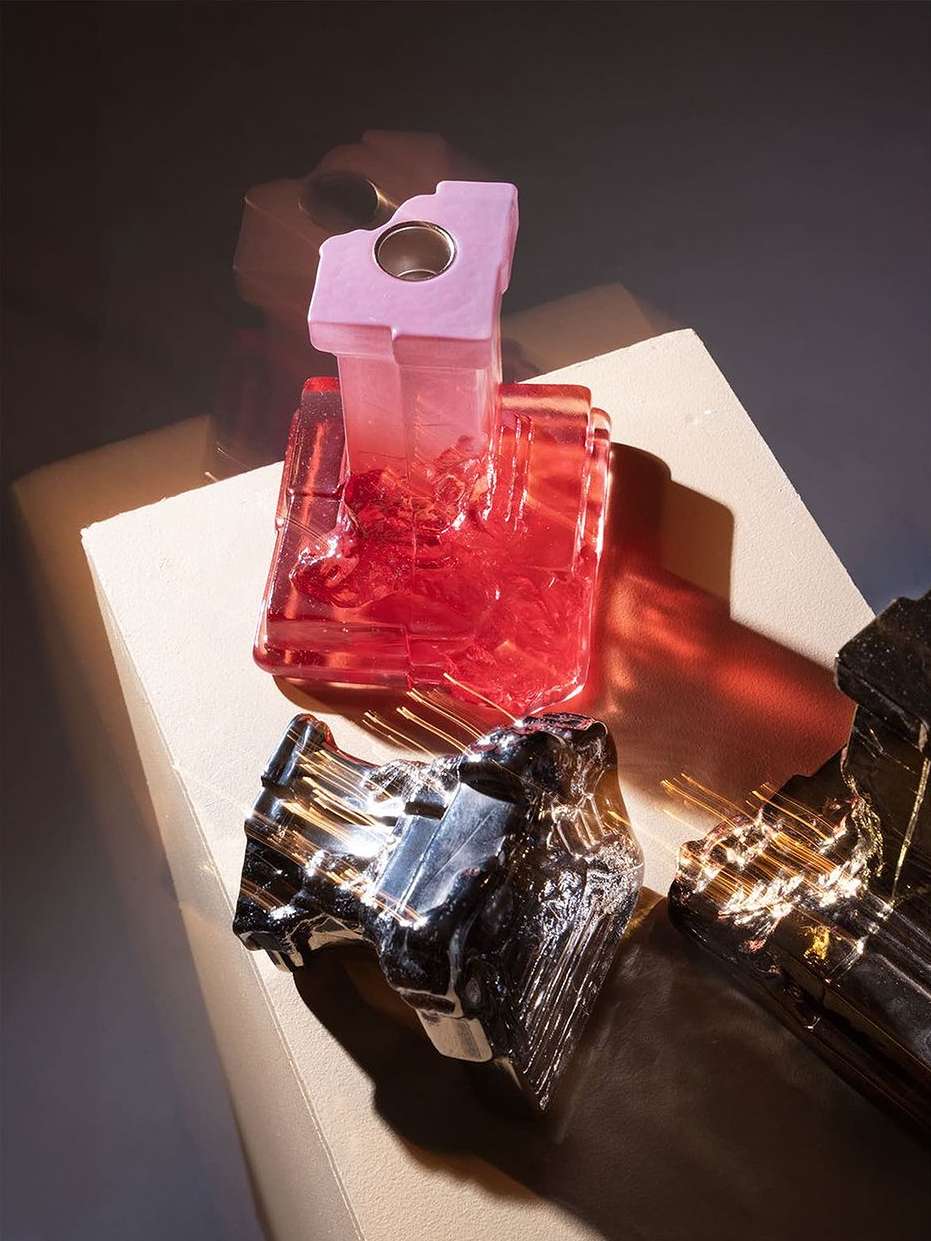 Three decorative glass objects in shades of pink, silver and black are displayed on a light brown surface, with a dark blurred background.