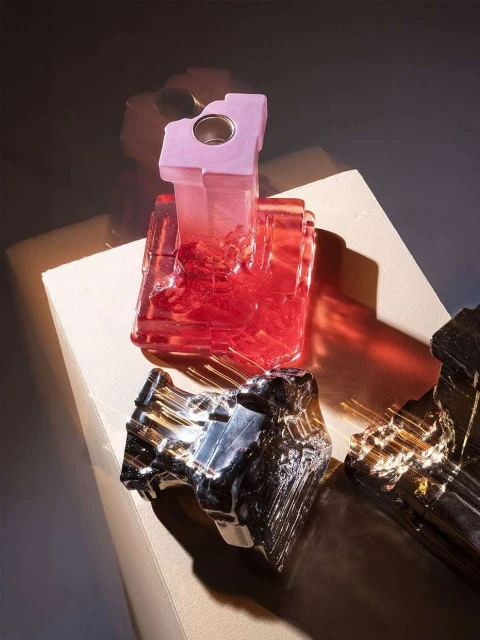 Three decorative glass objects in shades of pink, silver and black are displayed on a light brown surface, with a dark blurred background.