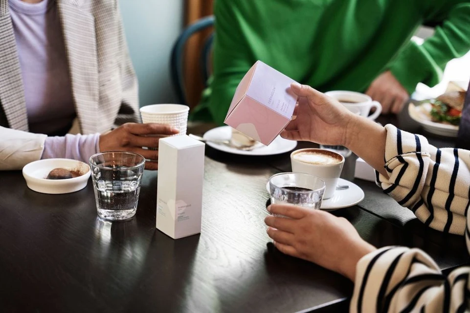 Three people at a table in a cafe or restaurant with coffee cups, drinking glasses, and product packaging on the dark wood table.
