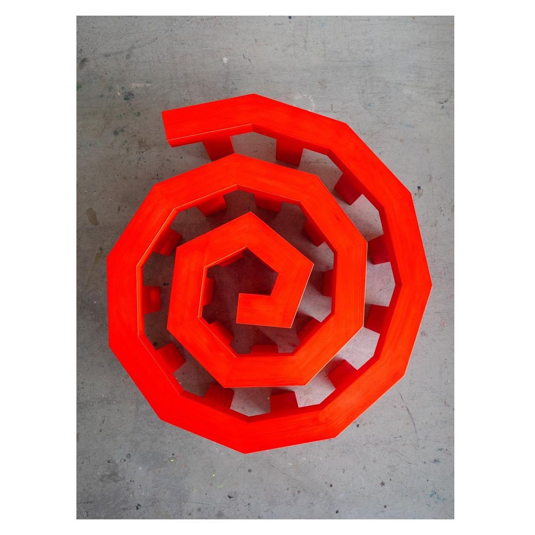 Overhead shot of an orange-red spiral structure with a hollow center, resting on a gray concrete surface, evoking a sense of modern art.