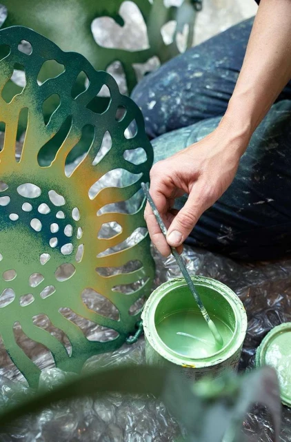 Person painting green on a metal lattice object with a paintbrush next to an open paint can in a studio setting with natural light.