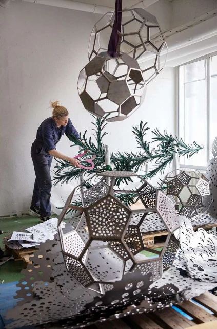 A woman using pink scissors to arrange green branches in large, geometric metal spheres inside a white-walled industrial space with natural light.