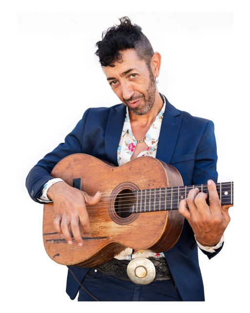 Man playing a worn acoustic guitar, wearing a floral shirt and navy blazer, with a white background. Musical expression.