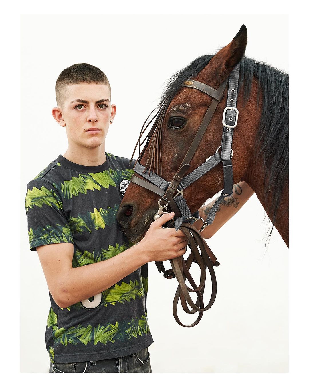 Teen poses with a brown horse in a studio. The horse has a bridle. The teen wears a patterned shirt, and they both face forward.