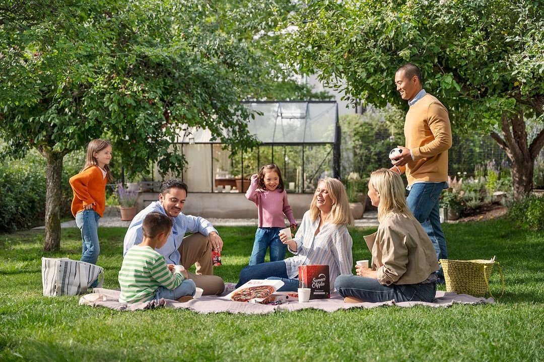 Family enjoys a picnic on a blanket in a grassy backyard under the shade of trees near a glass-enclosed structure during the day.
