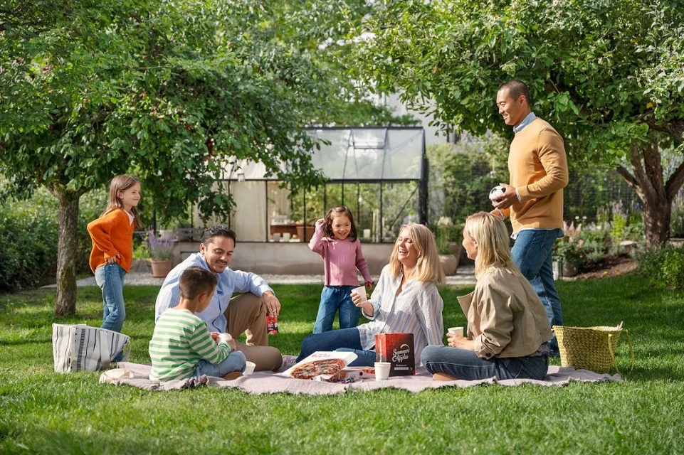 Family enjoys a picnic on a blanket in a grassy backyard under the shade of trees near a glass-enclosed structure during the day.