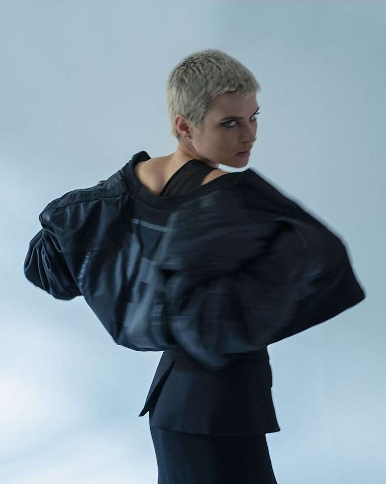 Stylish adult female fashion model posing in a photo studio, wearing a modern black skirt suit with a unique jacket design.