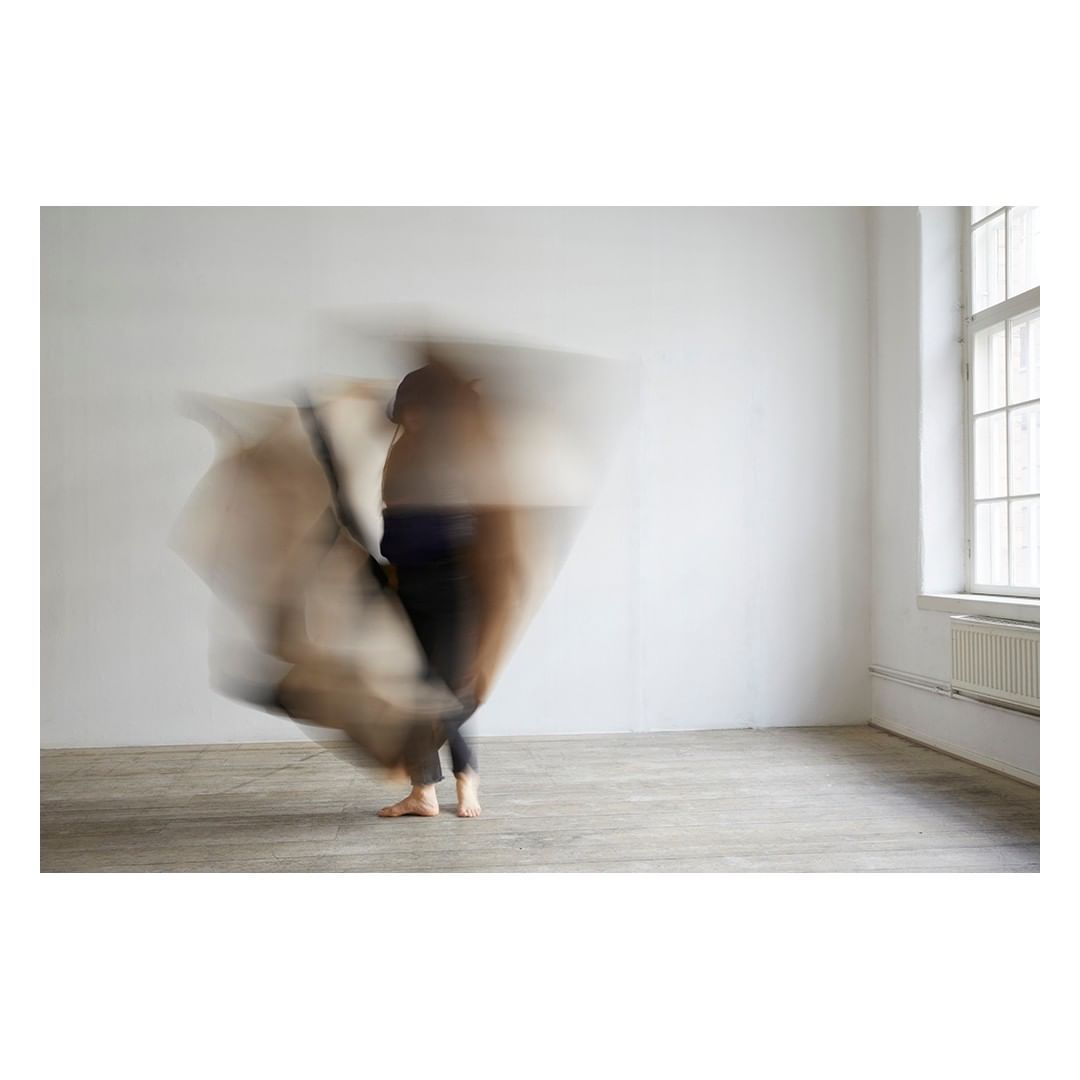 Motion blur photograph of a person in dark clothing, possibly dancing, inside a room with a white wall and window.