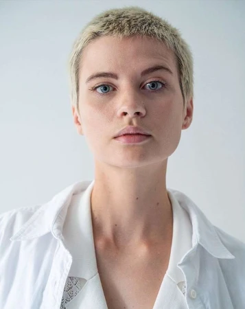 Close-up portrait of a woman with short blonde buzzcut hair wearing a white collared shirt against a pale seamless background.