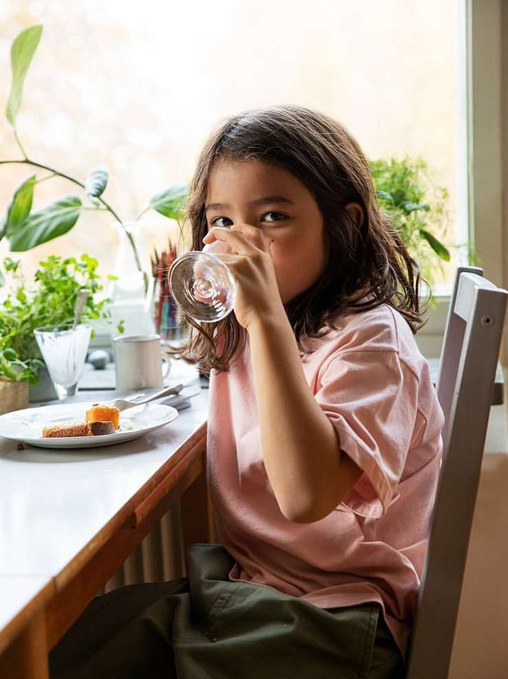 Young girl drinking water at a table near a window with plants, wearing a pink shirt and olive green pants, casual indoor setting.