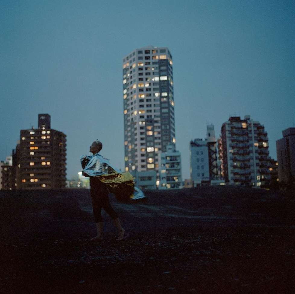Person in a reflective blanket stands on a dark surface with apartment buildings and night sky in the background at dusk.