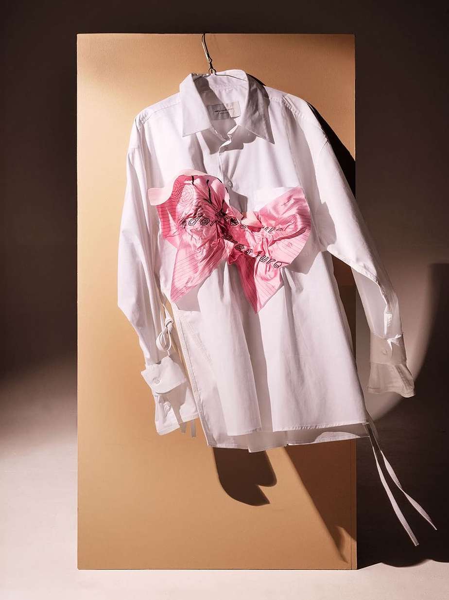 Fashion still life of a white collared shirt with a large pink ribbon hanging against a beige backdrop, studio lighting.