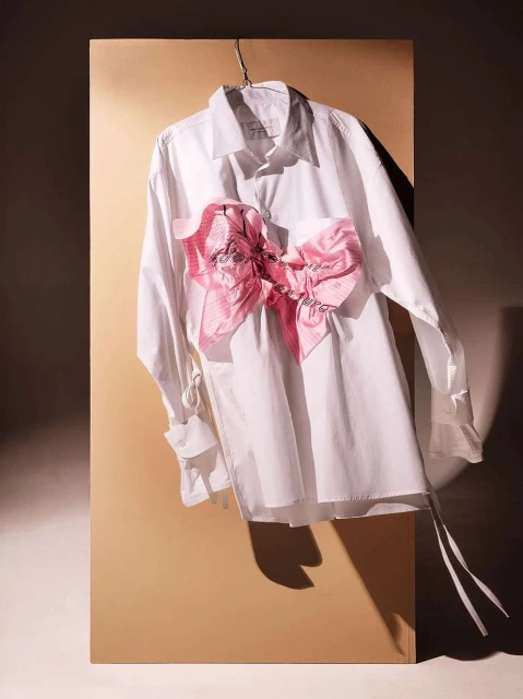 Fashion still life of a white collared shirt with a large pink ribbon hanging against a beige backdrop, studio lighting.
